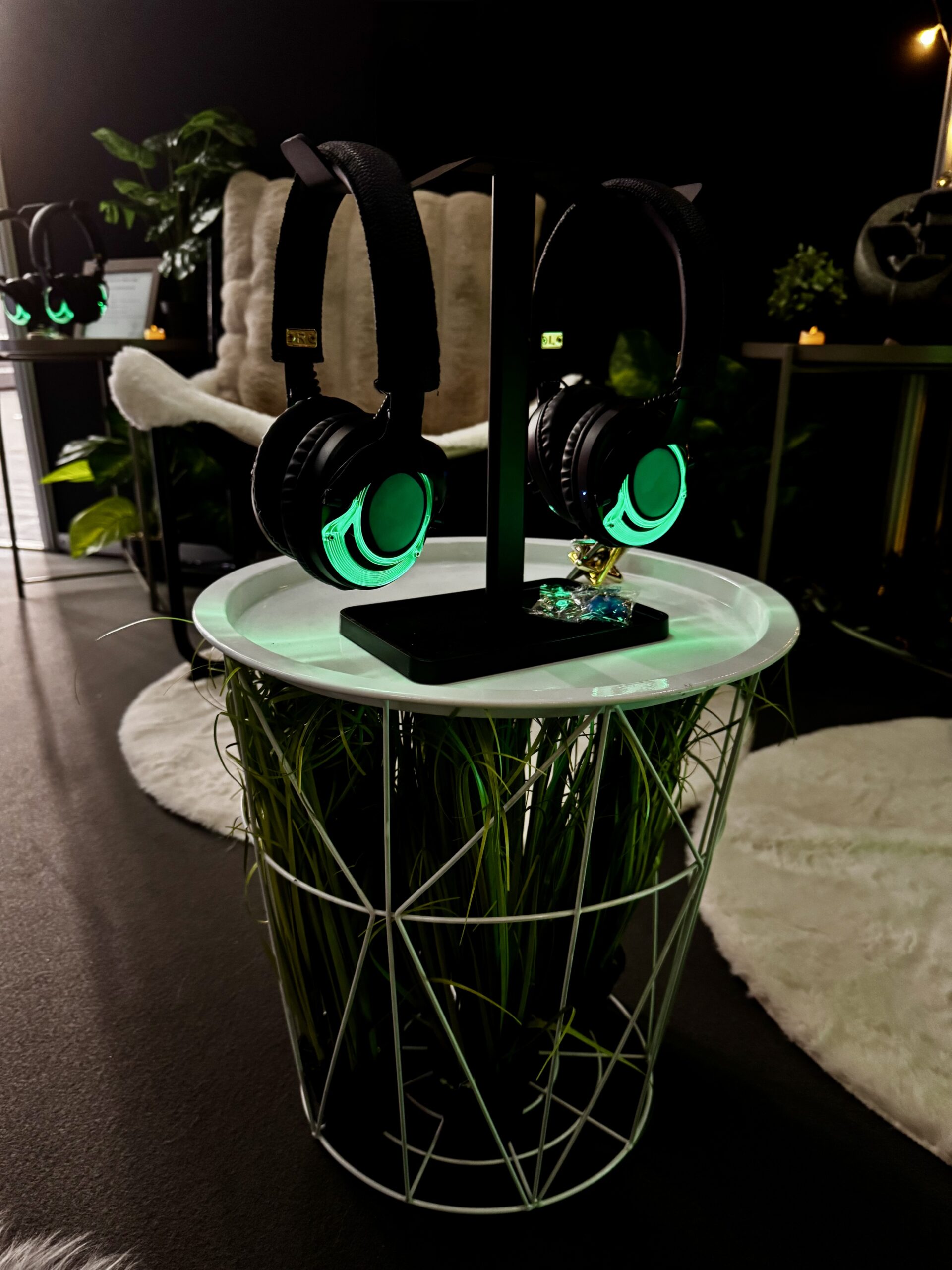 Four pairs of green-glowing headphones on white stand with neon accents, fur chair, plants, and basket nearby in dark booth