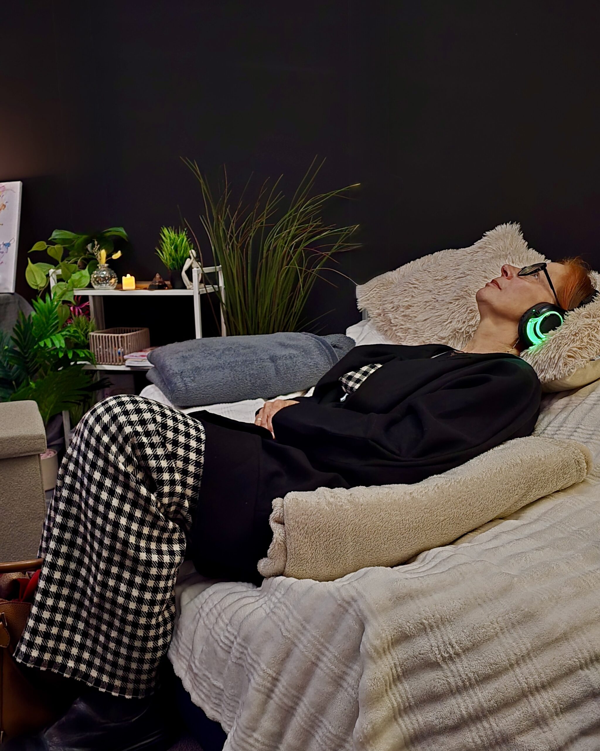 Attendee wearing headphones, lying on wide chair with pillows, surrounded by plants, lamps, fairy lights, and neon in Calm Nest sensory lounge.