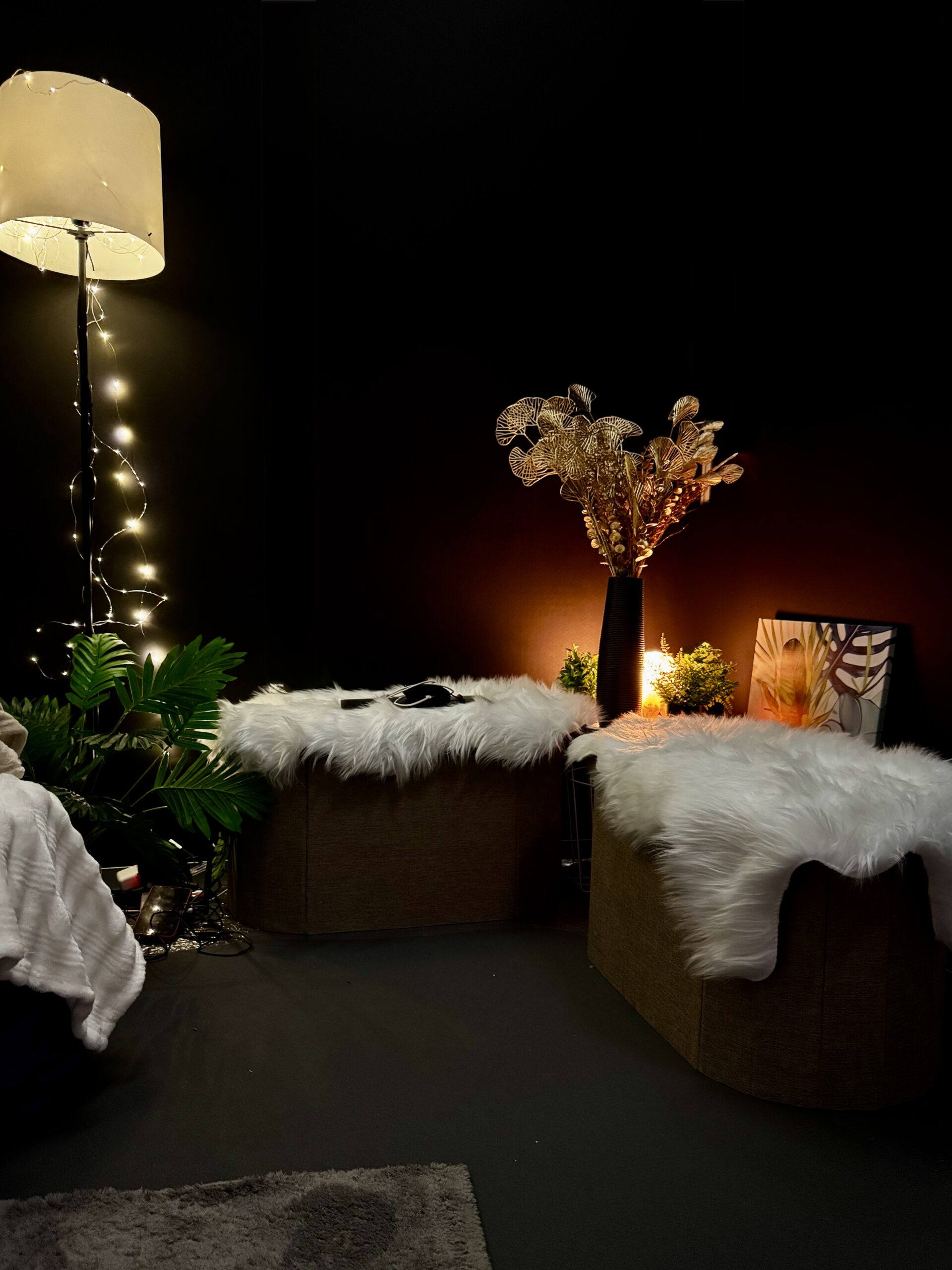 Low-lit Calm Nest corner with fur footstools, potted plants, candles, lamps, dried flowers, and woven stools on fluffy rugs for event calm zones.