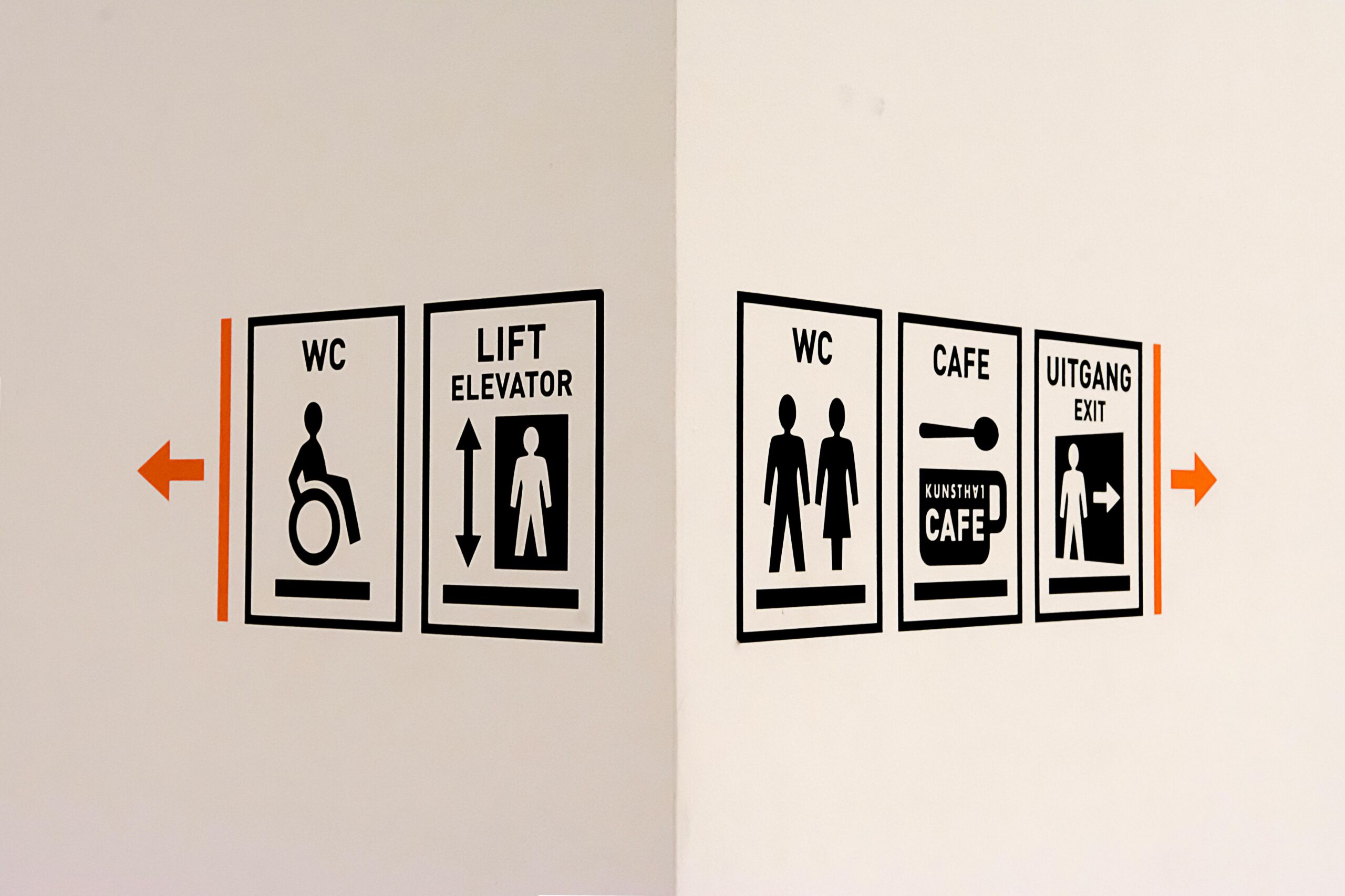 Clear black-and-white wayfinding signs with orange directional arrows showing WC, lift, café and exit at a public venue, representing best practice in accessible and neuroinclusive signage design
