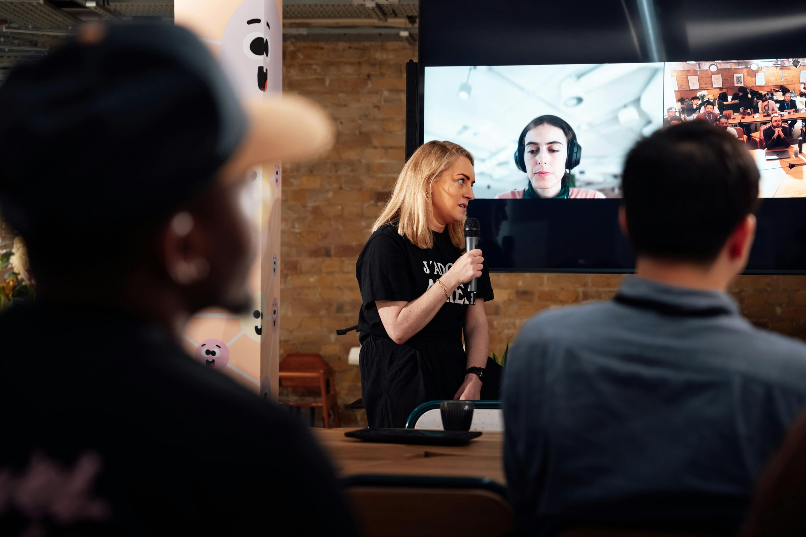 A woman speaking into a microphone at a hybrid event with a video screen showing remote participants, representing inclusive and accessible conference design