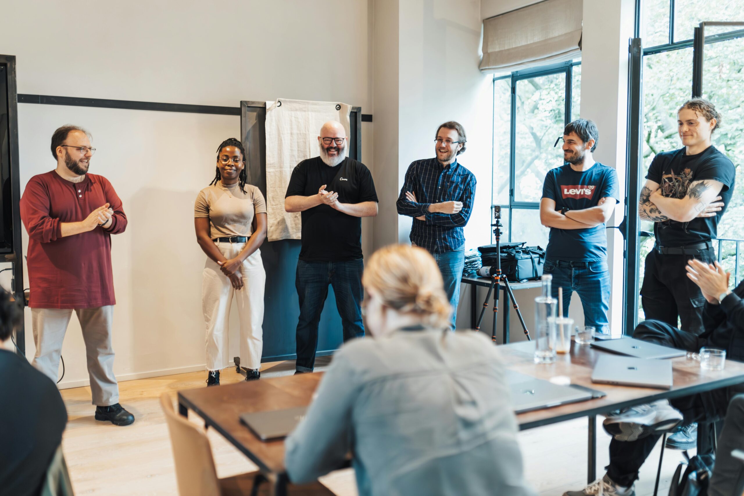 A diverse group of people smiling and standing at a bright, casual workshop space, representing inclusive team events and neuroinclusive workplace training facilitated by Calm Nest Collective